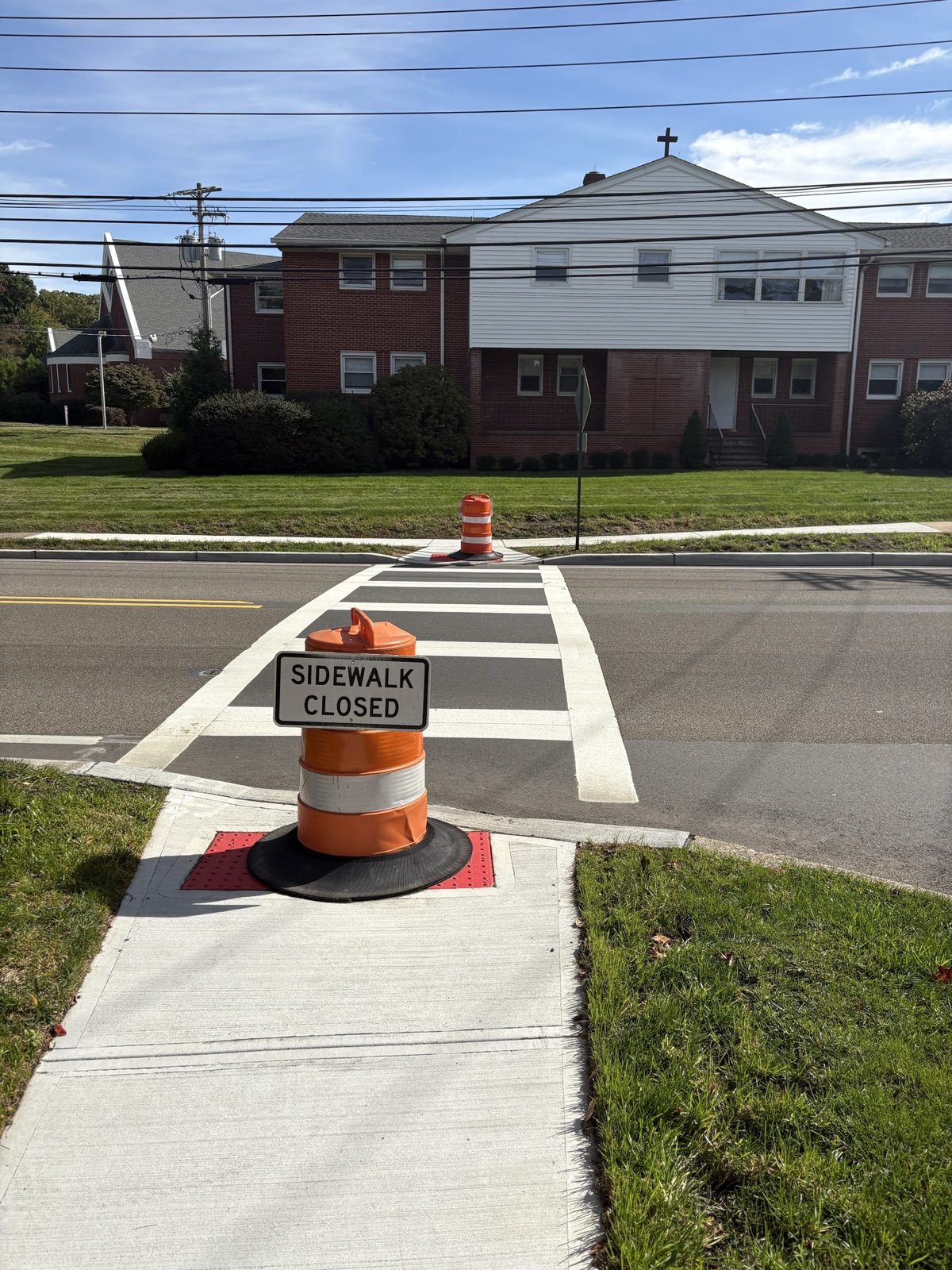 An orange traffic barrel with a "Sidewalk Closed" sign blocks a new curb ramp at the painted crosswalk across Wyckoff Avenue at Spring Meadow Drive, with a second barrel on the far side of the road.