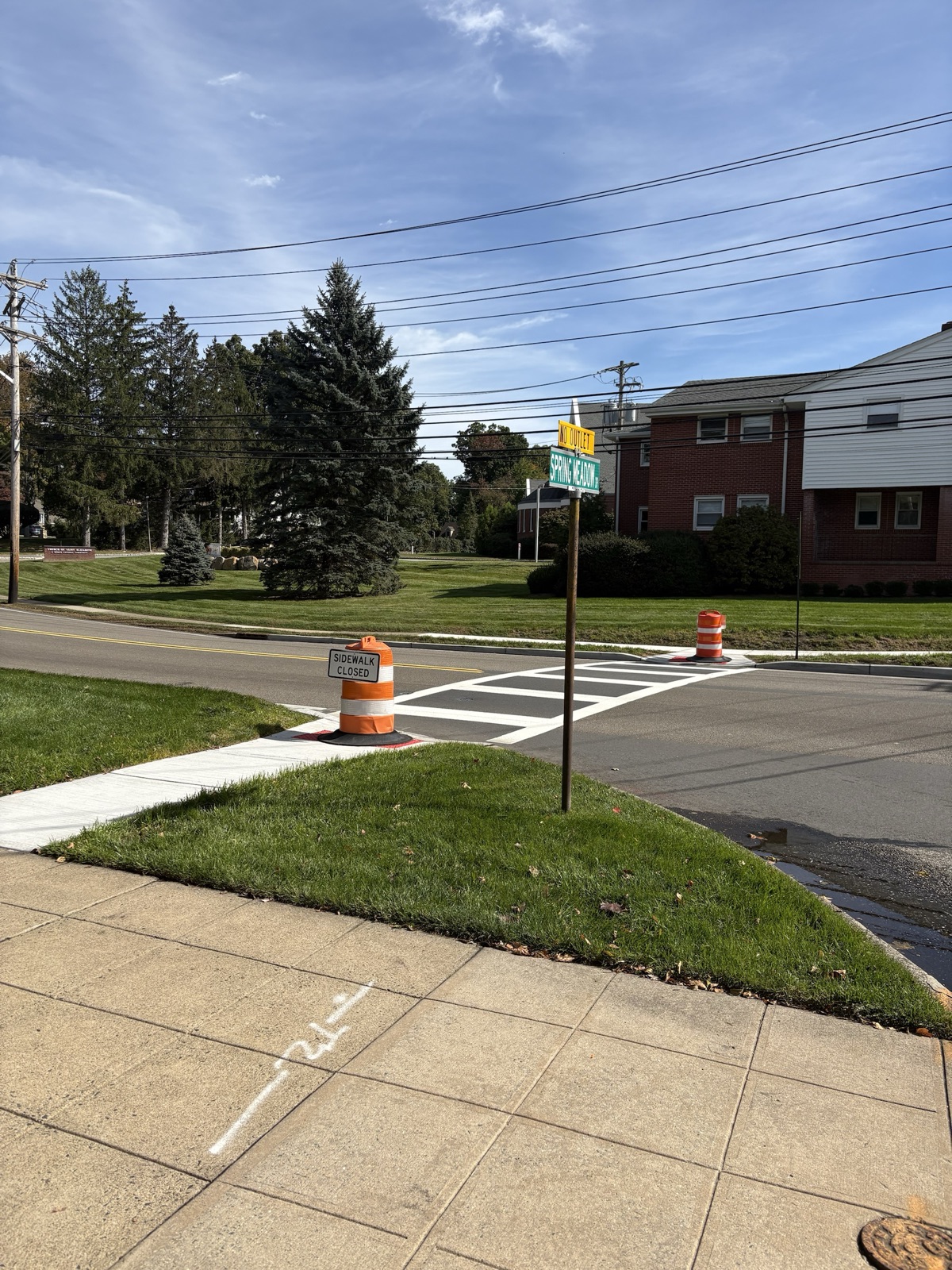 View from the Spring Meadow Drive street sign toward the crosswalk across Wyckoff Avenue, with barrels at both ends.