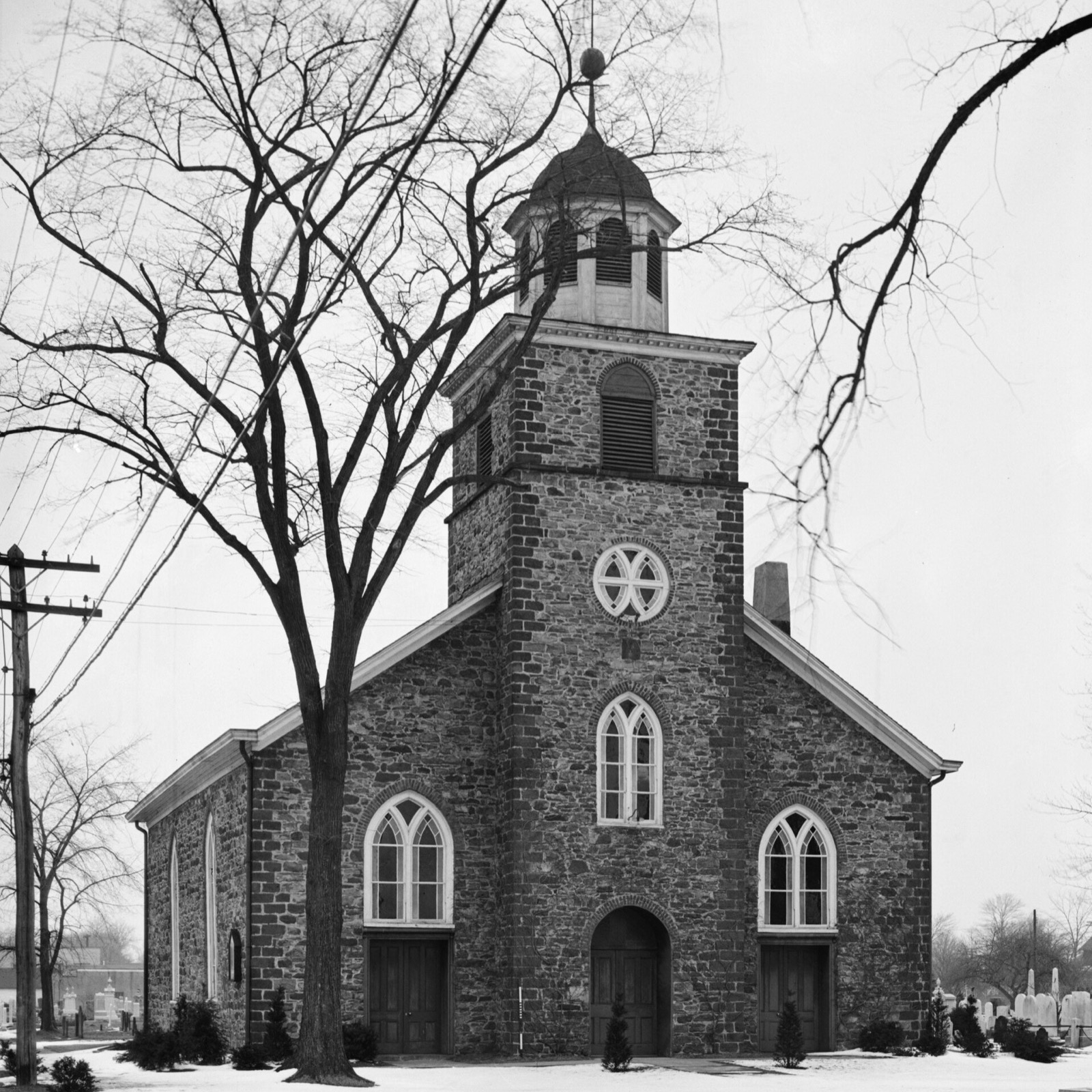Black-and-white 1937 photograph of the front of the fieldstone Wyckoff Reformed Church in snow, with a square bell tower and cupola, arched Gothic windows and doors, a bare tree in front, and gravestones visible at the right edge.