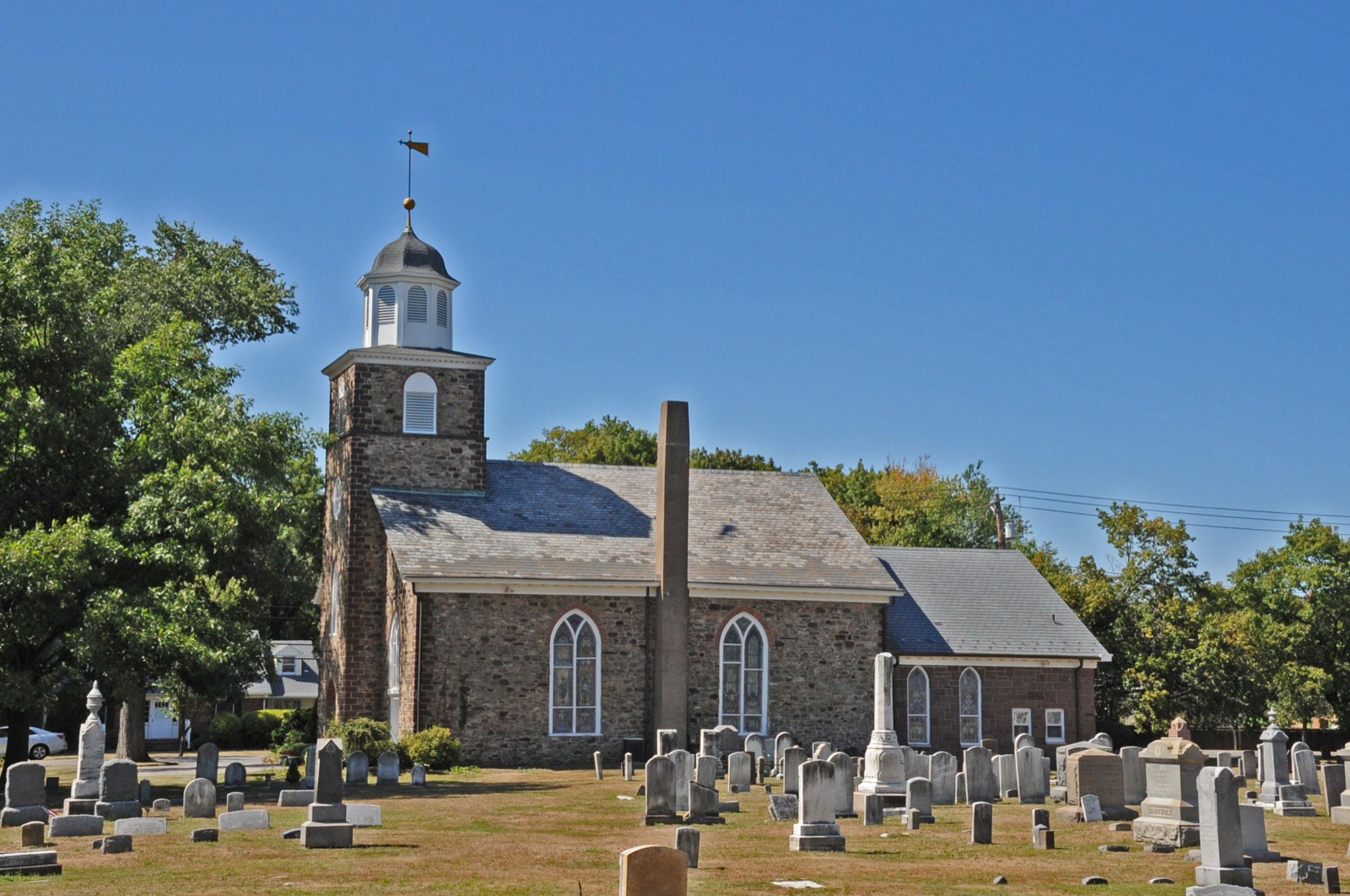 The fieldstone Wyckoff Reformed Church, with a square bell tower topped by a white cupola and weathervane, stands behind rows of gravestones and a tall obelisk monument in its cemetery under a clear blue sky.