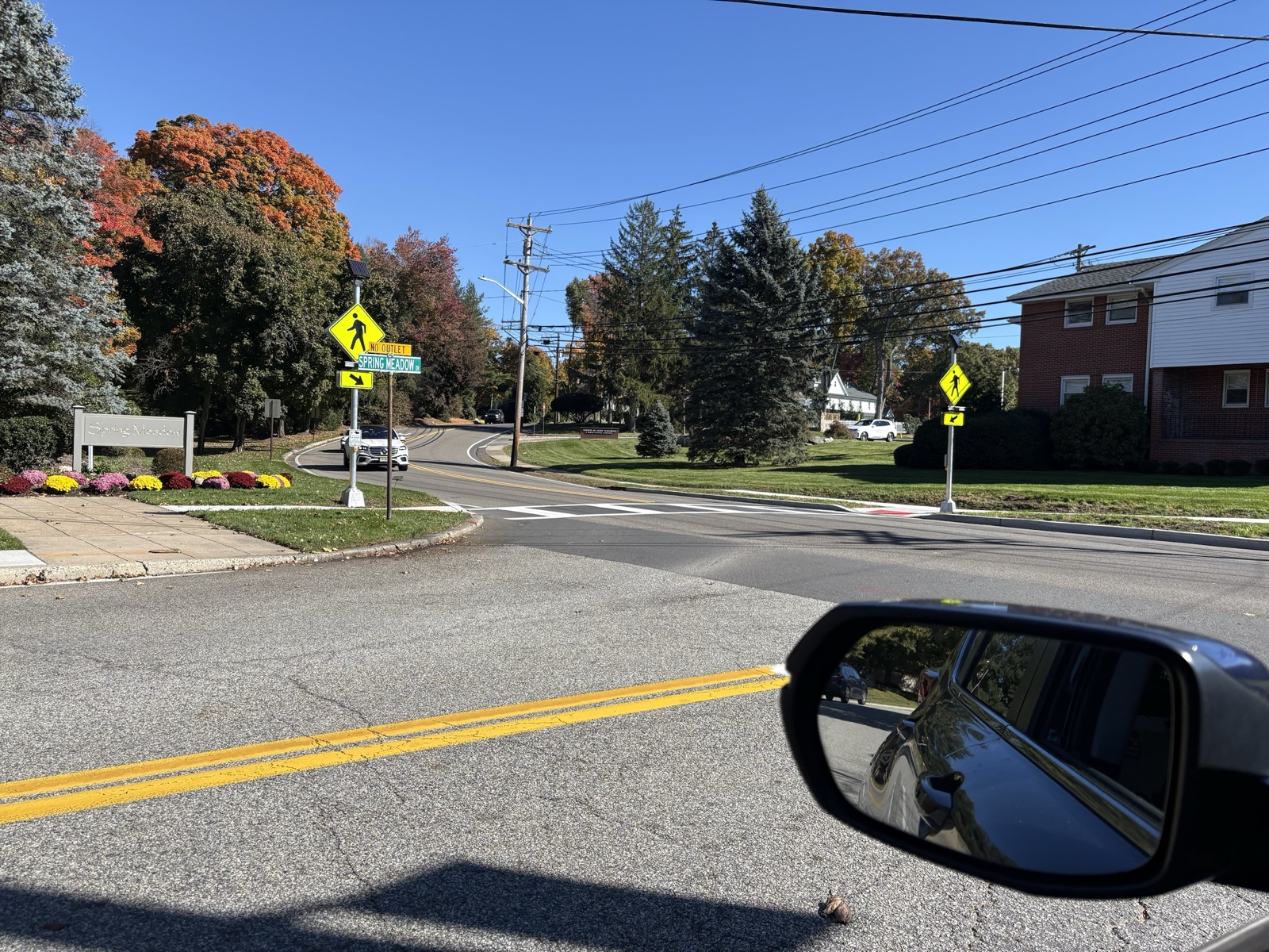 The crosswalk across Wyckoff Avenue at Spring Meadow Drive, seen from a car, with no barrels at the curb ramps and solar-topped beacon poles carrying yellow pedestrian-crossing signs at both ends. The Spring Meadow entrance sign and fall foliage are at left.